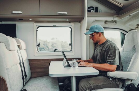 A man working in a Carado motorhome at the dinette with a table, tablet and window inside.