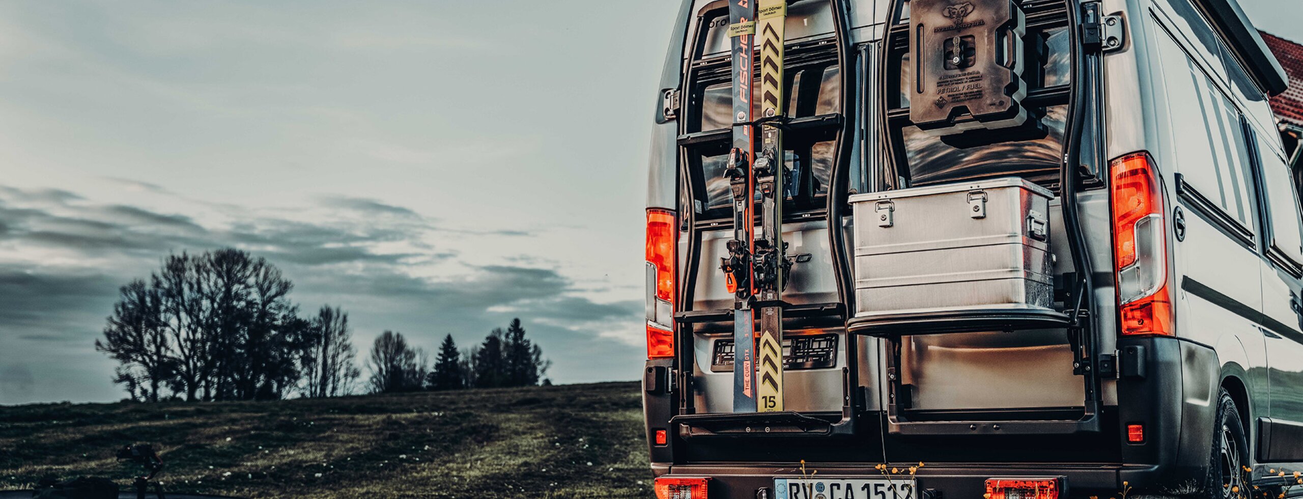 Vue arrière d’un camper van Carado avec porte-vélos, skis et porte arrière ouverte, avec coffre de rangement sur une prairie