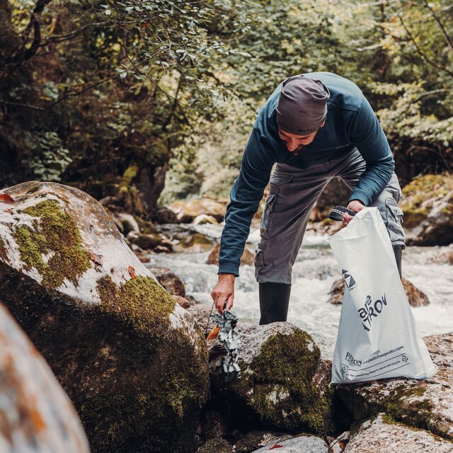 A man picks up litter among moss-covered rocks beside a stream in the forest