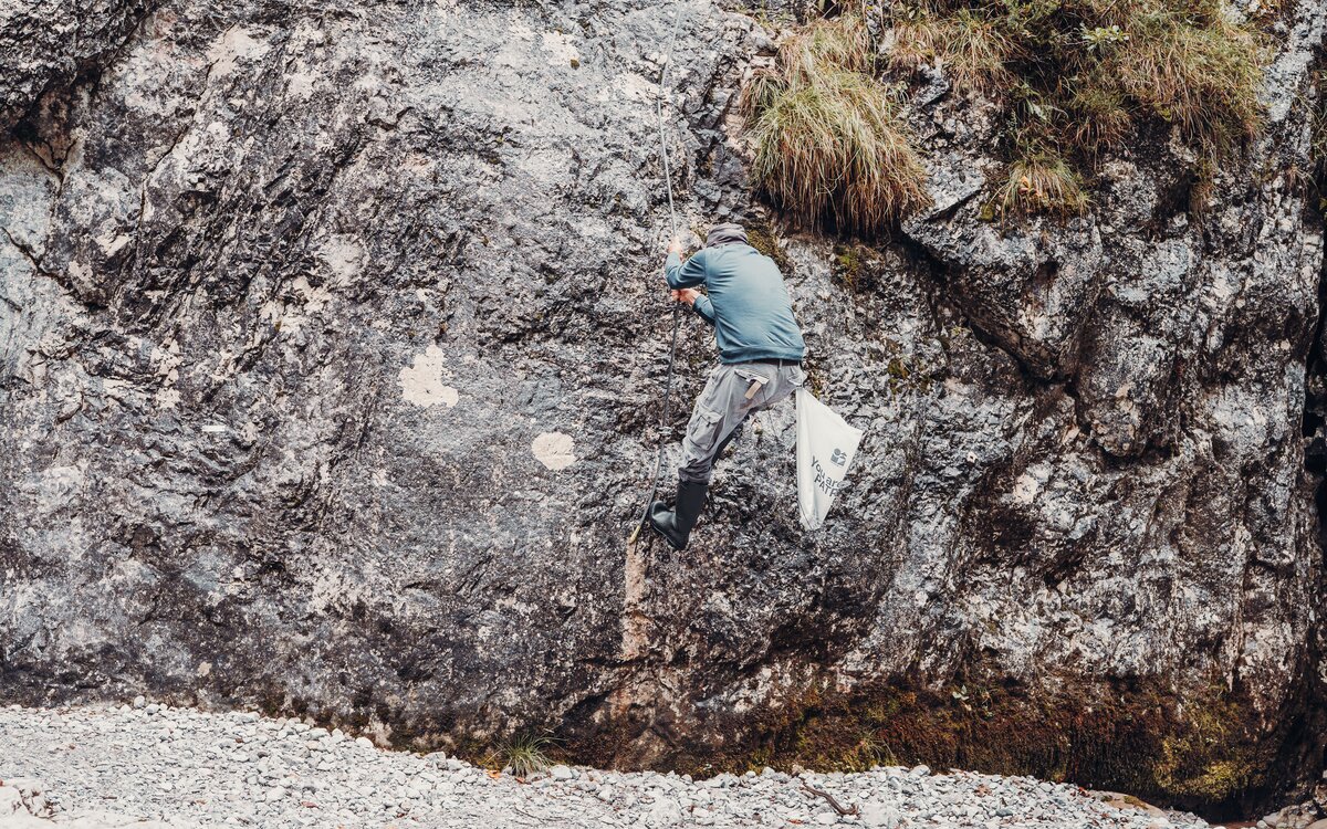 A person climbs a steep rock face with a rope above a rocky shore
