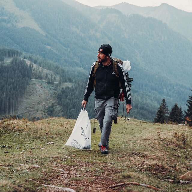 Backpacker with a rucksack and a rubbish bag on a mountain meadow in front of wooded slopes