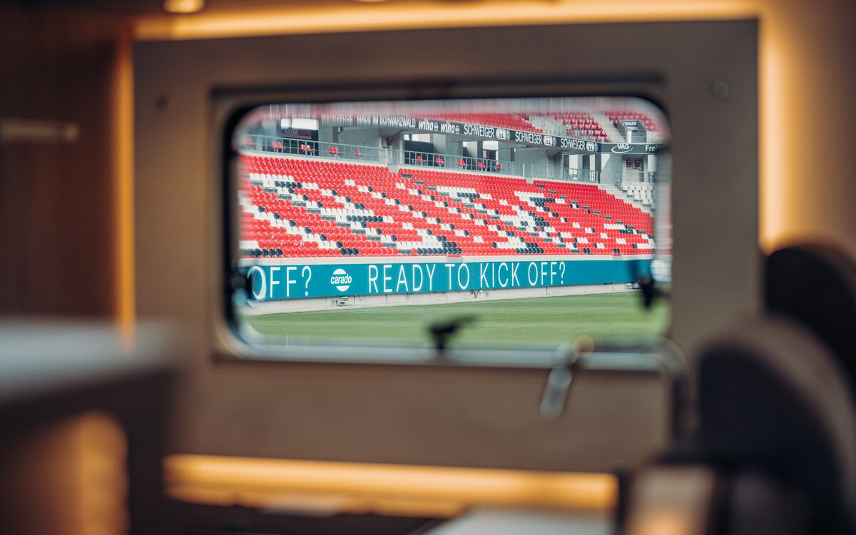 View from the window of a Carado motorhome to the grandstand of a stadium with red and white seats