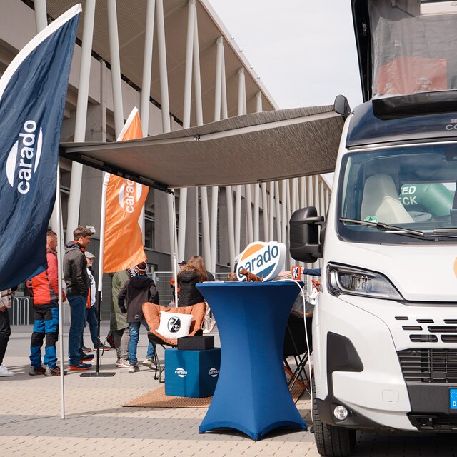 Carado motorhome with pop-up roof and awning at the exhibition grounds, people at the stand with Carado flags