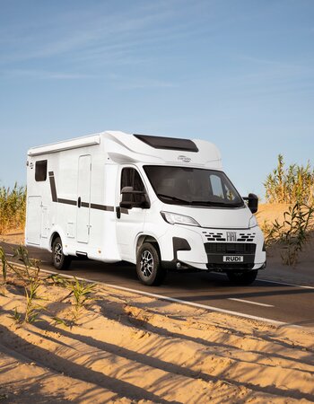 Carado motorhome in three-quarter view on a paved road between sand dunes and reeds