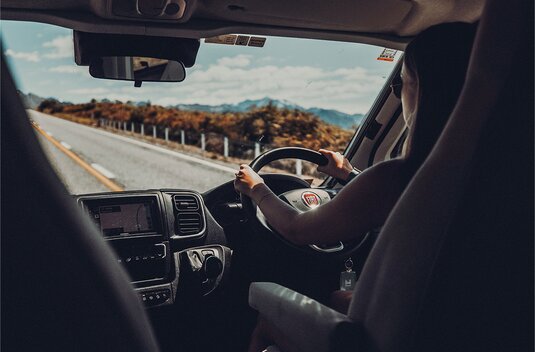Driver at the wheel of a Carado motorhome, looking through the windscreen at a country road and mountains