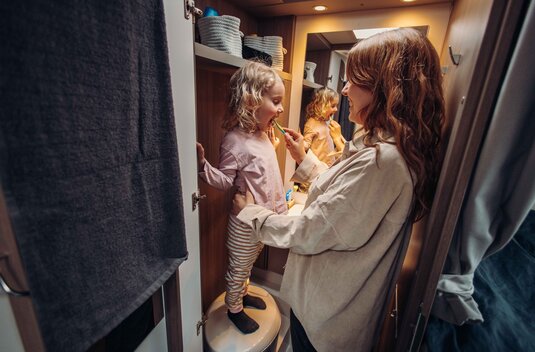 Une mère brosse les dents de son enfant dans la salle de bains d’un camping-car Carado, avec le miroir et les rangements en arrière-plan