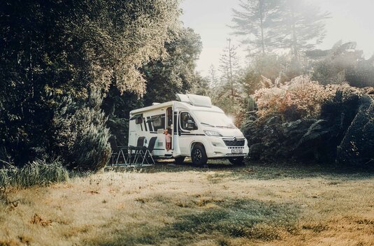 Camping-car Carado dans une clairière en forêt, un enfant dans l’embrasure de la porte ouverte, deux chaises de camping devant