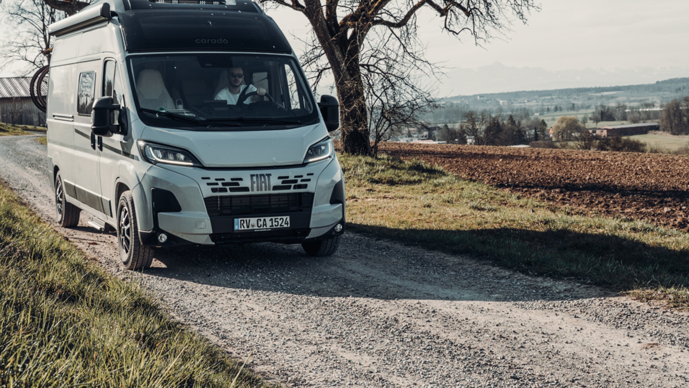 Carado motorhome on a gravel road in front of bare trees and fields, front view in a rural landscape