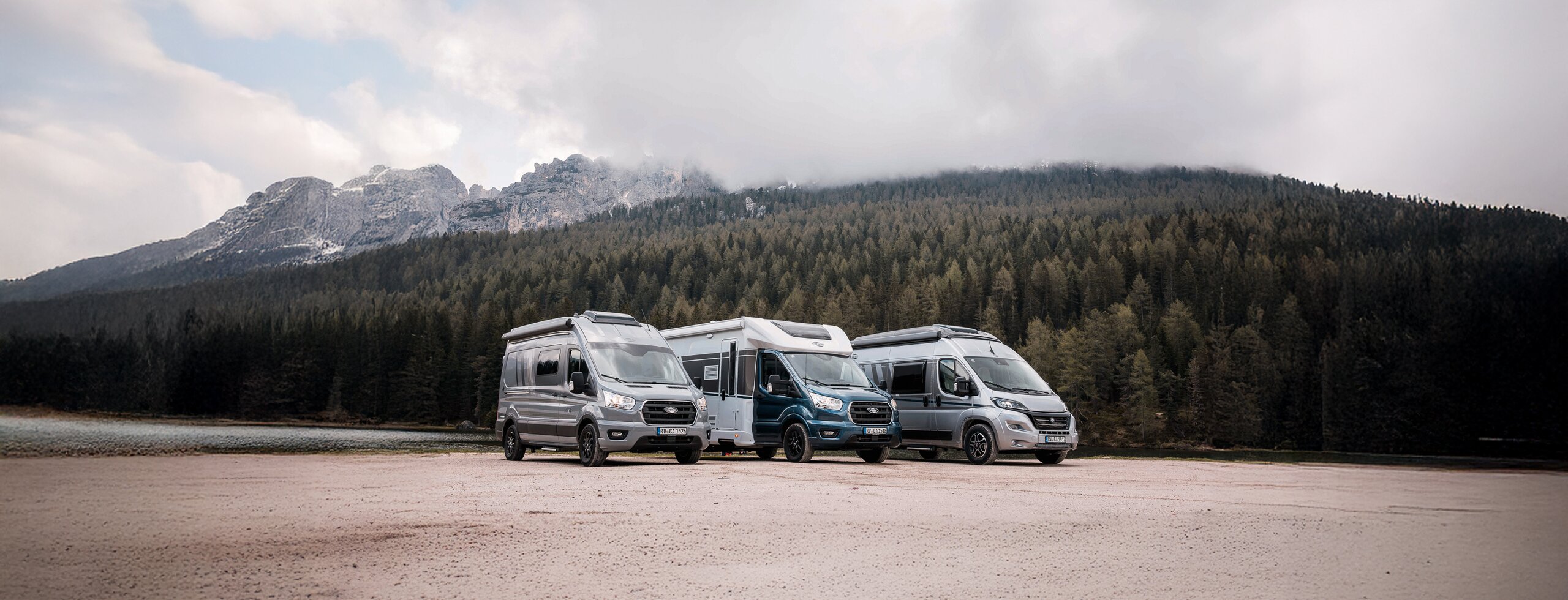Trois camping-cars et vans Carado sur un espace dégagé, avec forêt, lac et montagnes en arrière-plan