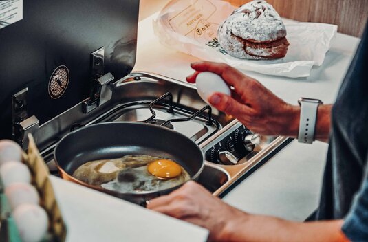 Cuisine de camping-car Carado avec plaque de cuisson à gaz, poêle avec œuf au plat et une main tenant un œuf