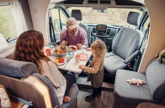 Famille à table dans le camping-car Carado, coin repas avec sièges avant pivotants et table dressée