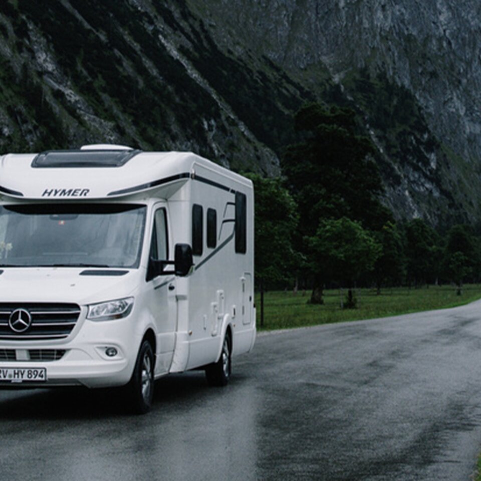 A white HYMER Tramp S is parked on a narrow, rain-soaked road in a mountainous landscape, with mountains, trees and meadows in the background.