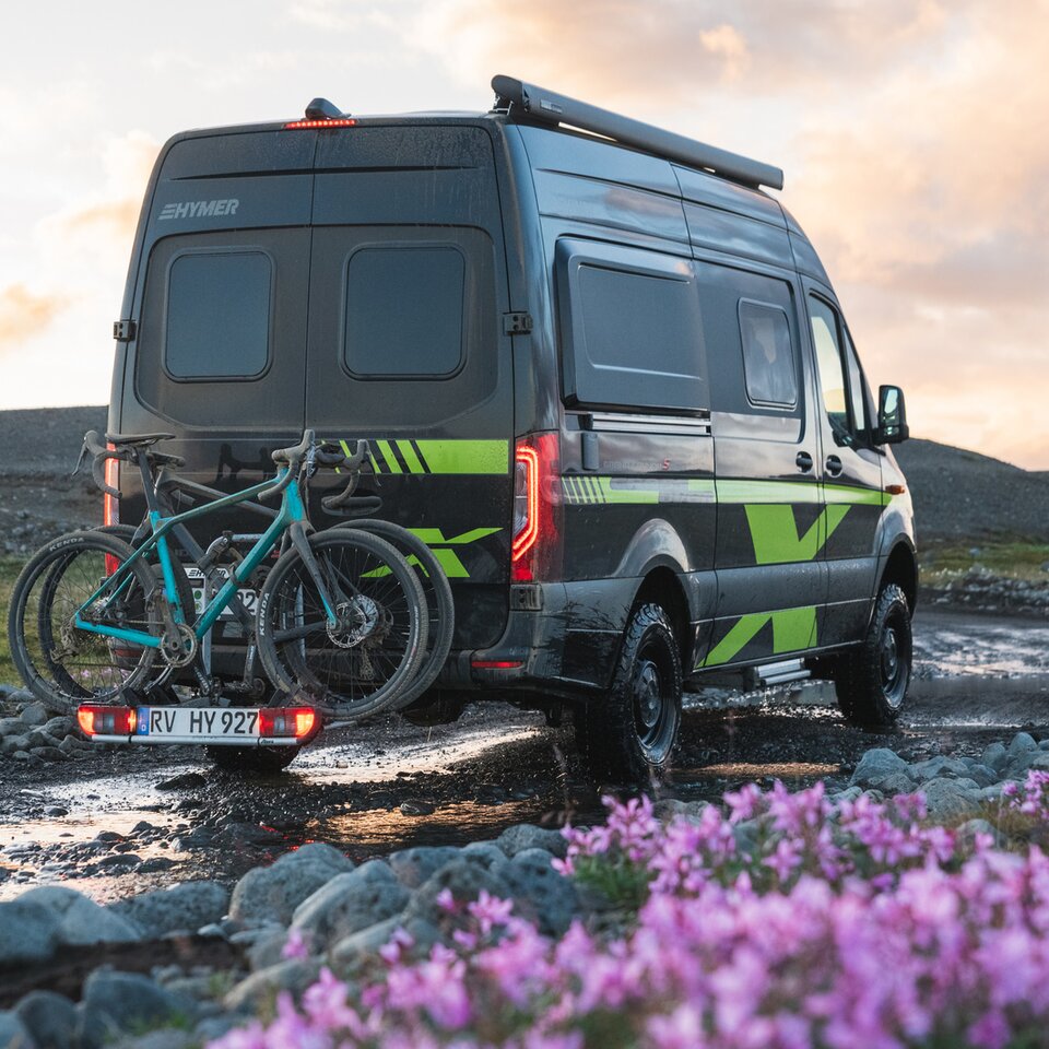 The Sprinter 4x4 motorhome HYMER Grand Canyon S CrossOver is parked on a muddy path with flowers and grass, with bikes on the bike rack.