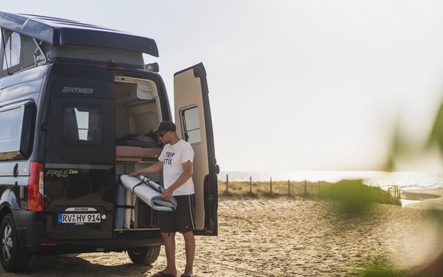 Mann tar en gjenstand gjennom bakdøren fra en mørk HYMER Campervan med åpent tak, parkert på stranden