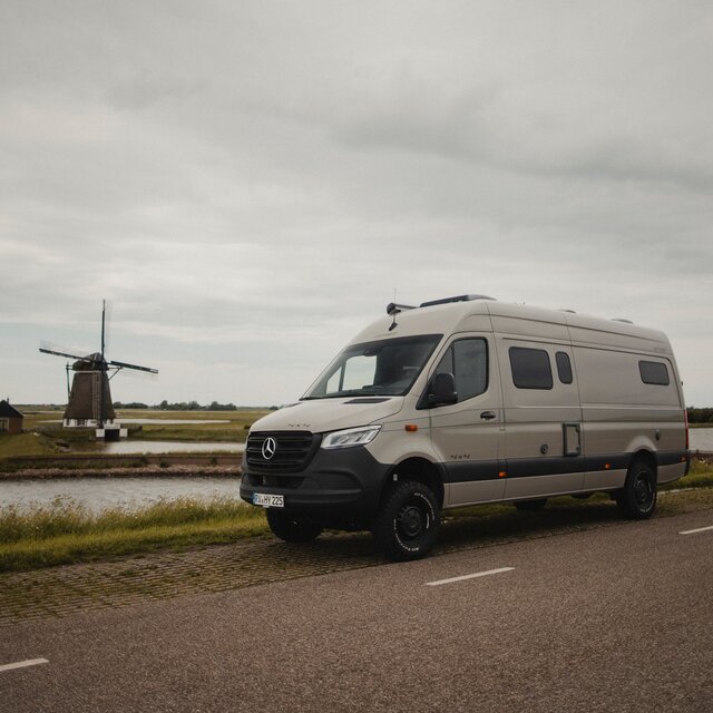Grey camper van stands sideways next to a road, behind it you can see a river and a small green island with a small windmill