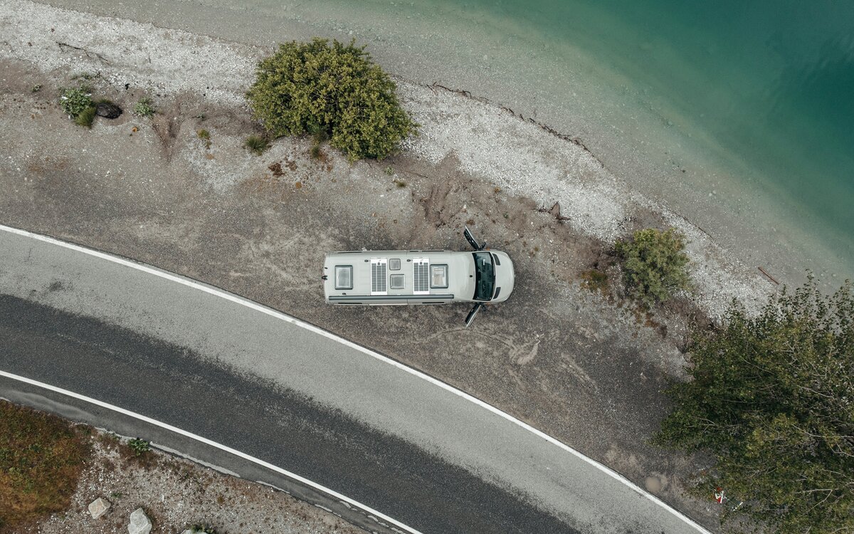 Grauer Camper Van ob oben, der auf Kies neben einem See am Ufer steht neben dem Kies gibt es eine kleine Straße und auf dem Kiesbett stehen drei grüne Bäume, die Türen des Camper Van sind geöffnet