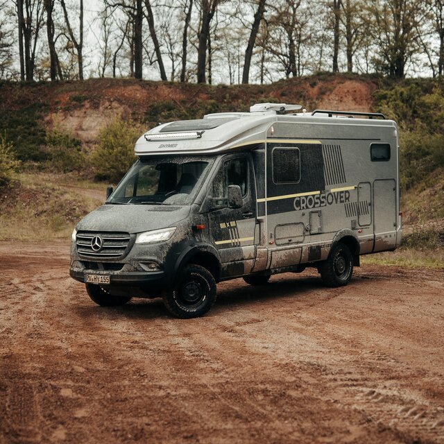 Semi-integrated 4x4 motorhome, seen from an angle in front, stands in an off-road park, trees and dirt mounds are visible in the background